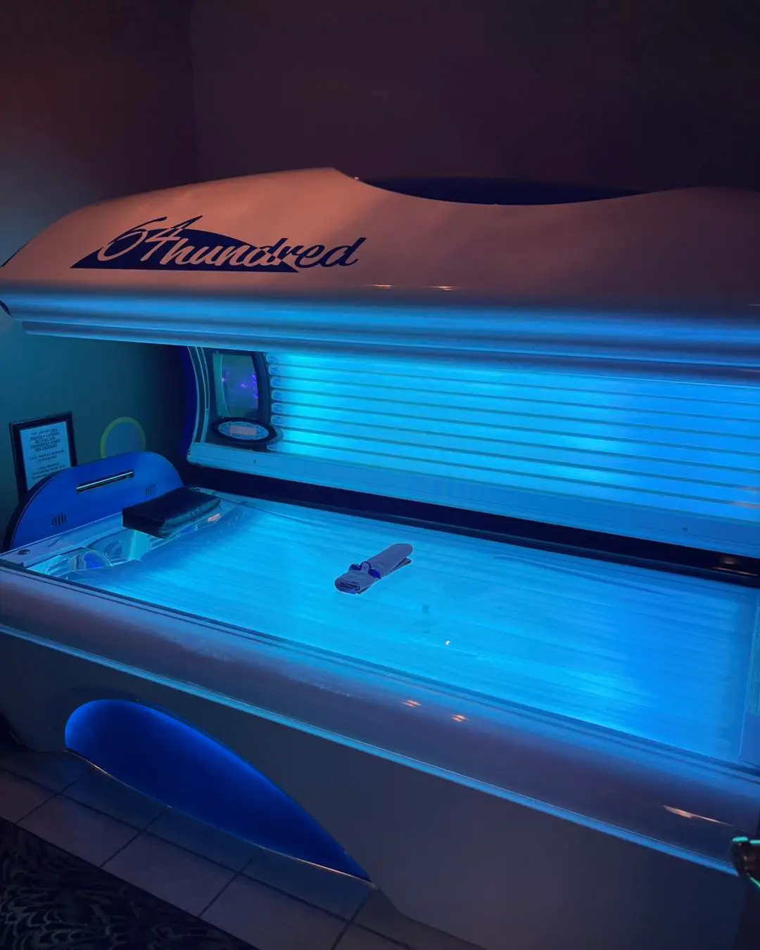 A white tanning bed with blue internal lighting is open in a dimly lit room, with the brand name "64hundred" visible.