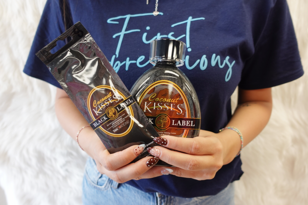 A person wearing a blue "First Impressions" shirt holds a packet and bottle of Coconut Kisses Black Label tanning lotion.