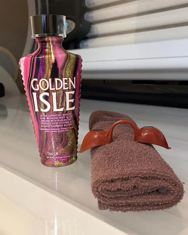A bottle of Golden Isle tanning lotion stands beside a folded brown towel and a pair of brown tanning goggles.