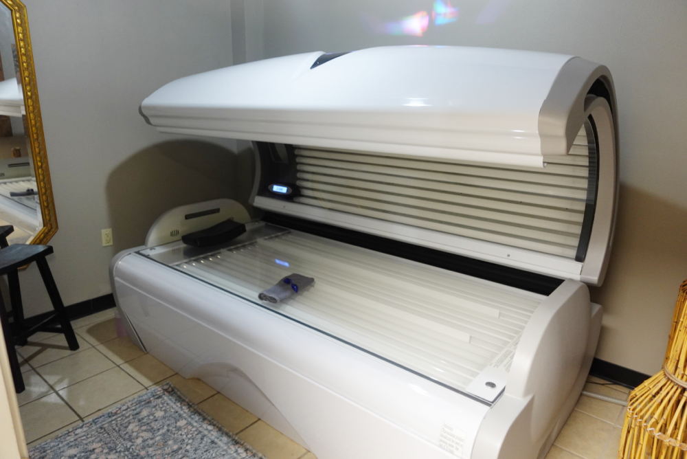 A tanning bed with its lid open, situated in a room with tiled floors and a gold-framed mirror.