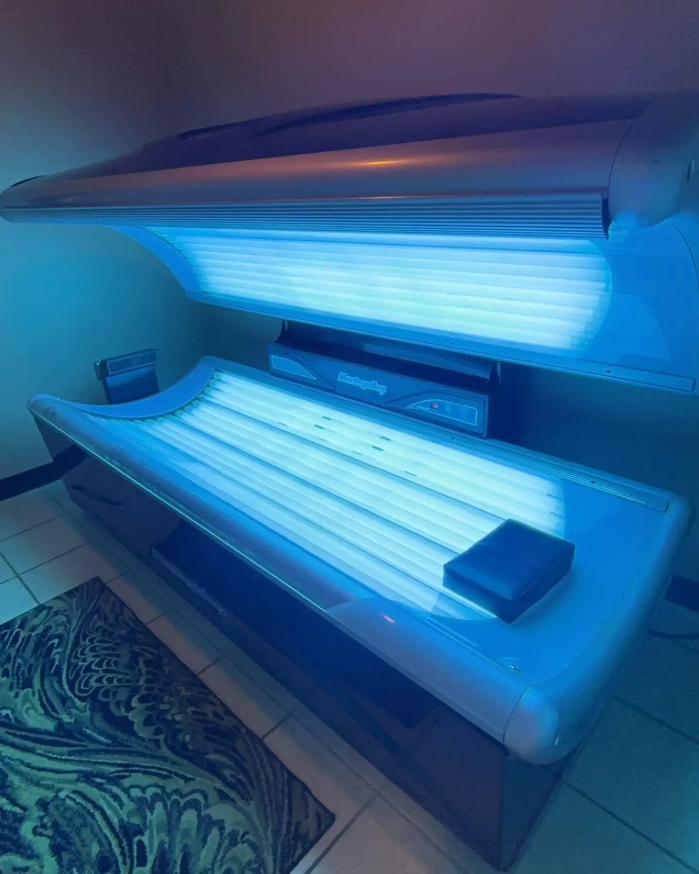 A glowing blue tanning bed stands open in a dimly lit, tiled room next to a patterned rug.